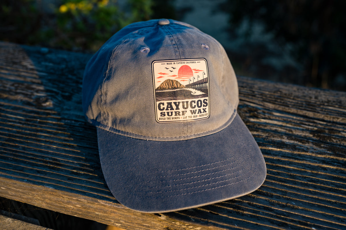 Blue Dougy Classic Dad hat resting on a weathered wooden surface outdoors, featuring a front logo that reads 'CAYUCOS SURF WAX' with a sunset and wave graphic.