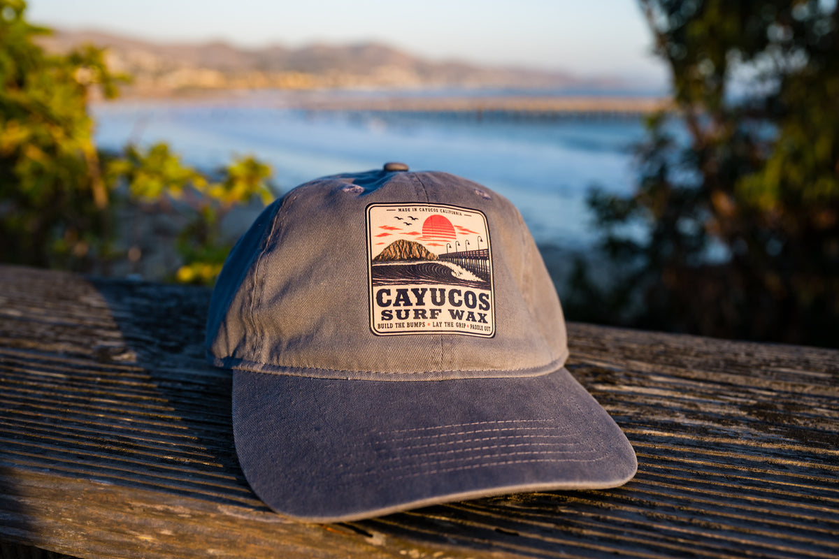 Blue Dougy Classic Dad hat resting on a weathered wooden surface outdoors, featuring a front logo that reads 'CAYUCOS SURF WAX' with a sunset and wave graphic.