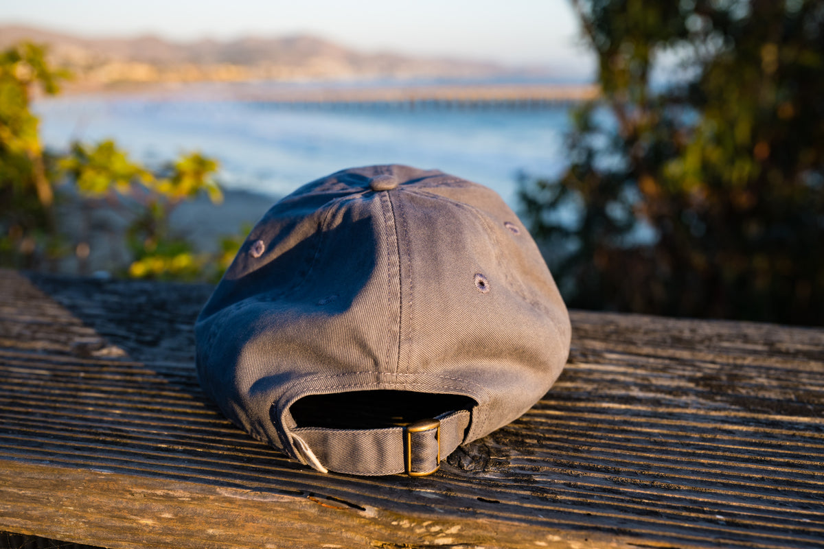 Back view of the Blue Dougy Classic Dad hat resting on a weathered wooden surface outdoors, featuring a front logo that reads 'CAYUCOS SURF WAX' with a sunset and wave graphic.