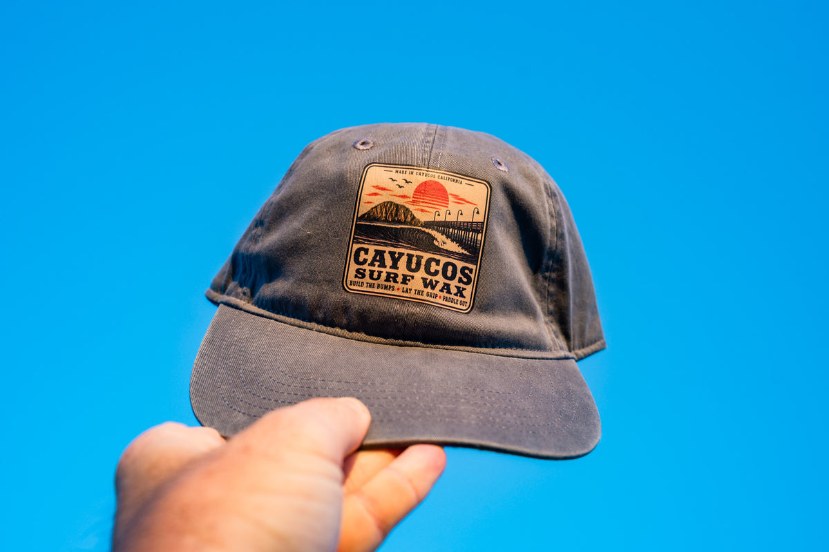 Hand holding the Blue Dougy Classic Dad hat, featuring a front logo that reads 'CAYUCOS SURF WAX' with a sunset and wave graphic.