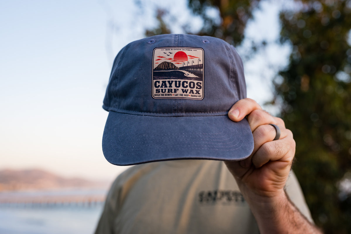 Blue Dougy Classic Dad hat featuring a front logo that reads 'CAYUCOS SURF WAX' with a sunset and wave graphic.